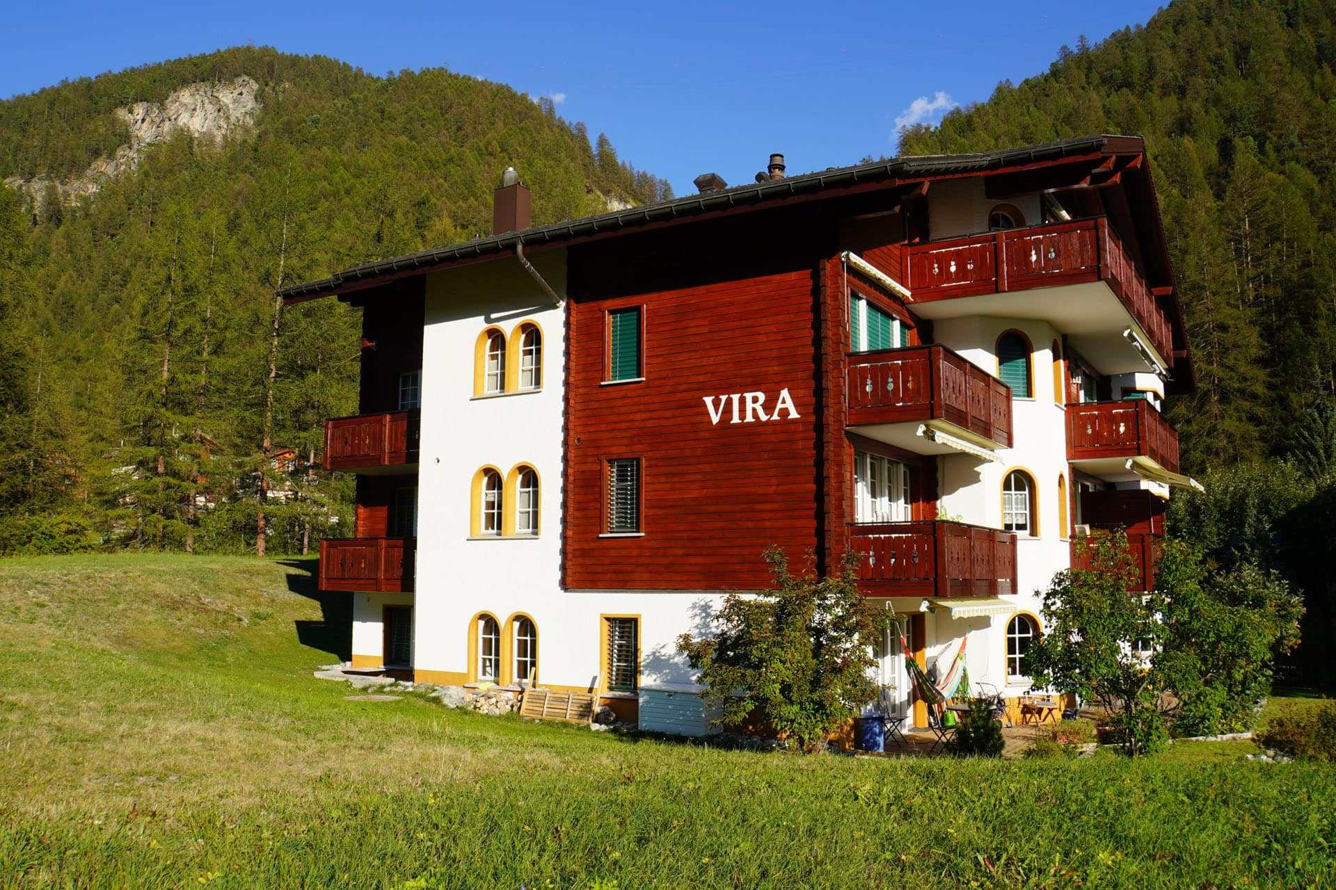 Haus Vira mit Holzbalkonen und dem Schriftzug VIRA, dahinter ein bewaldeter Hang